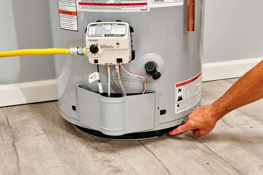 Hand pointing to water at the base of a gas water heater on a plank floor